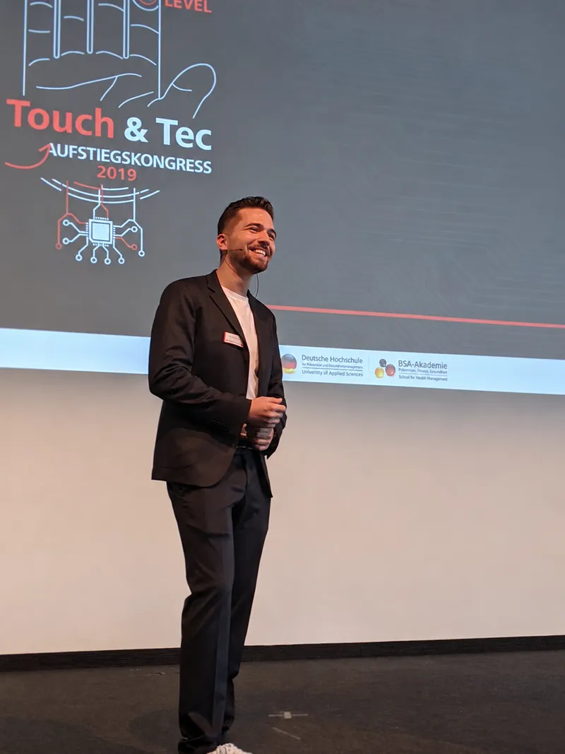 Frederik Neust lacht auf der Bühne des Touch & Tec Aufstiegskongresses 2019, auf der Leinwand die Logos der DHfPG und der BSA-Akademie