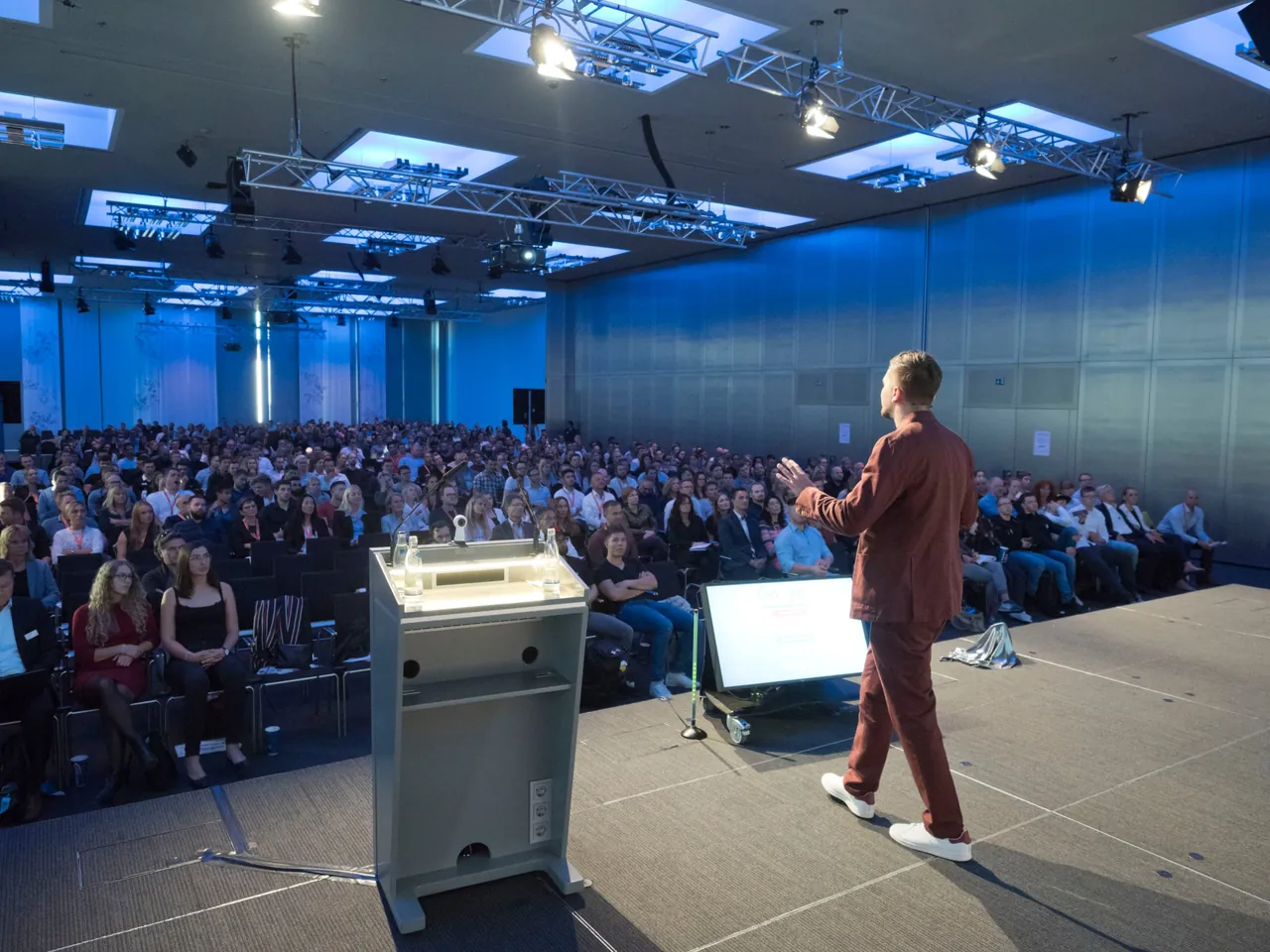 SEO-Speaker Frederik Neust im rotbraunen Anzug spricht vor einem voll besetzten Saal, von hinten fotografiert